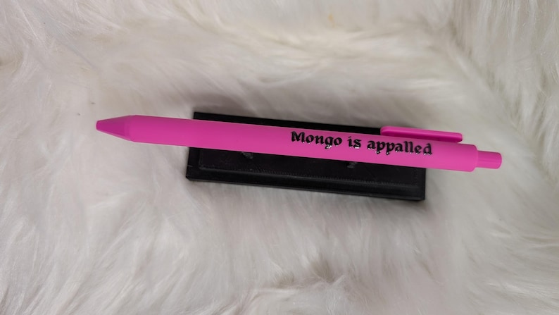 Puede incluir: Un bol&iacute;grafo retr&aacute;ctil de color rosa brillante con el texto en negro Mongo is appalled impreso en el cuerpo. El bol&iacute;grafo incluye un clip de bolsillo rosa a juego y est&aacute; colocado horizontalmente sobre un soporte negro.