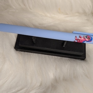 Puede incluir: Un bol&iacute;grafo retr&aacute;ctil de color azul claro con un peque&ntilde;o gr&aacute;fico de una criatura rosa y azul en el cuerpo, cerca del clip. El bol&iacute;grafo descansa horizontalmente sobre un soporte rectangular negro.