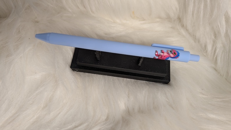Puede incluir: Un bol&iacute;grafo retr&aacute;ctil de color azul claro con un peque&ntilde;o gr&aacute;fico de una criatura rosa y azul en el cuerpo, cerca del clip. El bol&iacute;grafo descansa horizontalmente sobre un soporte rectangular negro.