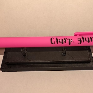 Puede incluir: Un bol&iacute;grafo retr&aacute;ctil de color rosa brillante con el texto negro "Glurp, glurp" impreso en el cuerpo. El bol&iacute;grafo incluye un clip de bolsillo rosa a juego y un mecanismo de bot&oacute;n en la parte superior, apoyado horizontalmente sobre un soporte negro.