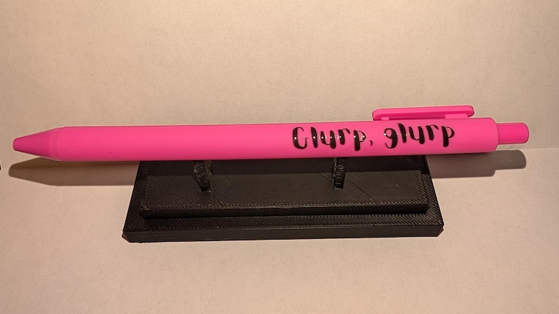 Puede incluir: Un bol&iacute;grafo retr&aacute;ctil de color rosa brillante con el texto negro "Glurp, glurp" impreso en el cuerpo. El bol&iacute;grafo incluye un clip de bolsillo rosa a juego y un mecanismo de bot&oacute;n en la parte superior, apoyado horizontalmente sobre un soporte negro.