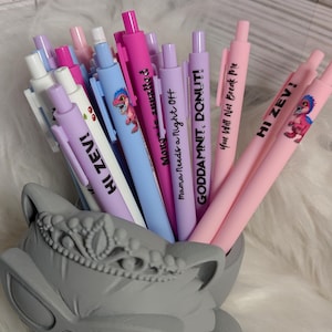 Puede incluir: Una colecci&oacute;n de bol&iacute;grafos retr&aacute;ctiles coloridos con varias frases y gr&aacute;ficos de dinosaurios, incluyendo textos como HI ZEV!, Mama Needs a Night Off, GODDAMNIT, DONUT! y You Will Not Break Me, en un soporte gris con forma de gato y tiara.