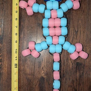 May include: A beaded lizard figurine, approximately 10 inches long, in light blue, pink, and dark blue. The lizard is made of cylindrical beads strung together with a silver-colored chain. The lizard is lying on a wooden surface.