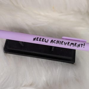 Puede incluir: Un bol&iacute;grafo retr&aacute;ctil de color morado claro con un clip y el texto negro NEEEW ACHIEVEMENT! impreso en el cuerpo. El bol&iacute;grafo descansa horizontalmente sobre un peque&ntilde;o soporte rectangular negro.