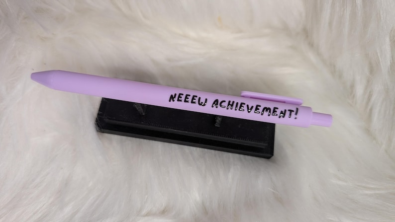 Puede incluir: Un bol&iacute;grafo retr&aacute;ctil de color morado claro con un clip y el texto negro NEEEW ACHIEVEMENT! impreso en el cuerpo. El bol&iacute;grafo descansa horizontalmente sobre un peque&ntilde;o soporte rectangular negro.