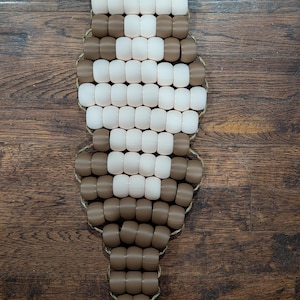 May include: A beaded, decorative item in a vertical orientation. The piece is made of brown, black, and white beads strung together. The beads are arranged in a pattern, with a rope-like border. The item is displayed on a wooden surface.