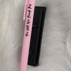 Puede incluir: Un bol&iacute;grafo retr&aacute;ctil de color rosa claro con un clip de bolsillo y el texto impreso en negro "You Will Not Break Me" que recorre verticalmente el cuerpo, colocado junto a un objeto rectangular negro.