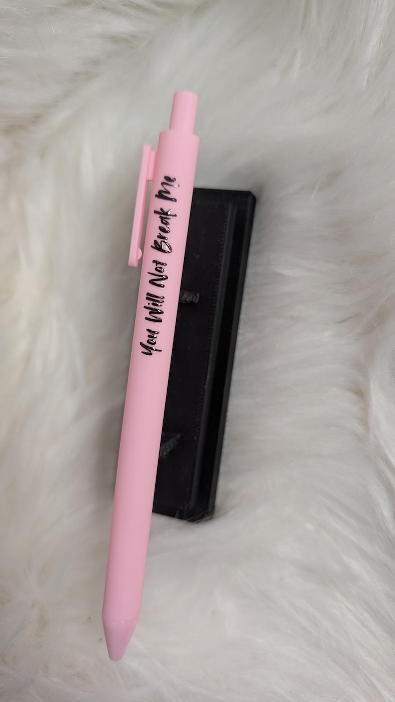 Puede incluir: Un bol&iacute;grafo retr&aacute;ctil de color rosa claro con un clip de bolsillo y el texto impreso en negro "You Will Not Break Me" que recorre verticalmente el cuerpo, colocado junto a un objeto rectangular negro.