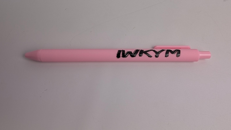 Puede incluir: Un bol&iacute;grafo retr&aacute;ctil de color rosa claro con acabado mate y clip de bolsillo. El texto IWKYM est&aacute; impreso en letras may&uacute;sculas negras y negritas a lo largo del cuerpo del bol&iacute;grafo, colocado horizontalmente cerca del extremo superior.