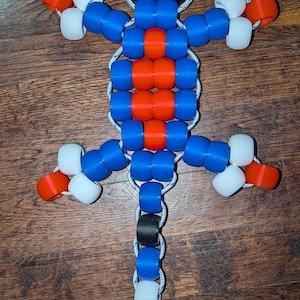 Puede incluir: Una figura de lagarto hecha con cuentas de plástico azul, naranja, blanco y negro ensartadas en un cordón blanco. El cuerpo presenta un patrón central de cuentas naranjas rodeadas de azul, con detalles en blanco y naranja en las extremidades.