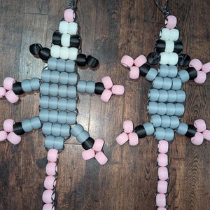 May include: Two lizard-shaped ornaments crafted from plastic pony beads in shades of gray, black, white, and light pink. Each figure features a long tail, four legs, and a loop at the top for hanging, strung together with a dark gray cord.