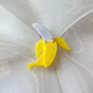 Puede incluir: Un par de pendientes amarillos en forma de plátano con puntas blancas. Los pendientes están hechos de plástico y tienen un acabado brillante.