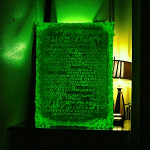 May include: A glowing green rectangular plaque with an ancient script. The edges have a textured, rough appearance. The text is in a dark green color, and the background is a lighter green. The plaque is illuminated by a green light.