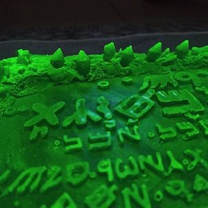 May include: A close-up of a green, textured surface with raised symbols and shapes. Several crystal-like structures are arranged along the top edge. The symbols and crystals are illuminated with a bright green light.