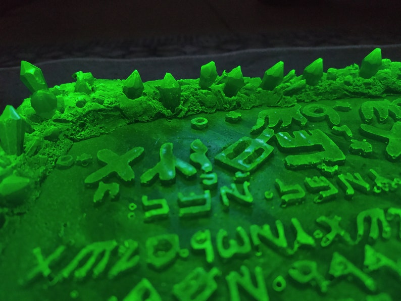 May include: A close-up of a green, textured surface with raised symbols and shapes. Several crystal-like structures are arranged along the top edge. The symbols and crystals are illuminated with a bright green light.