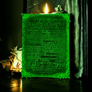 May include: A glowing green candle with ancient-looking text carved into its surface. The candle is rectangular with a textured, uneven edge. A lit flame sits atop the candle, casting a warm glow. The background is dark.