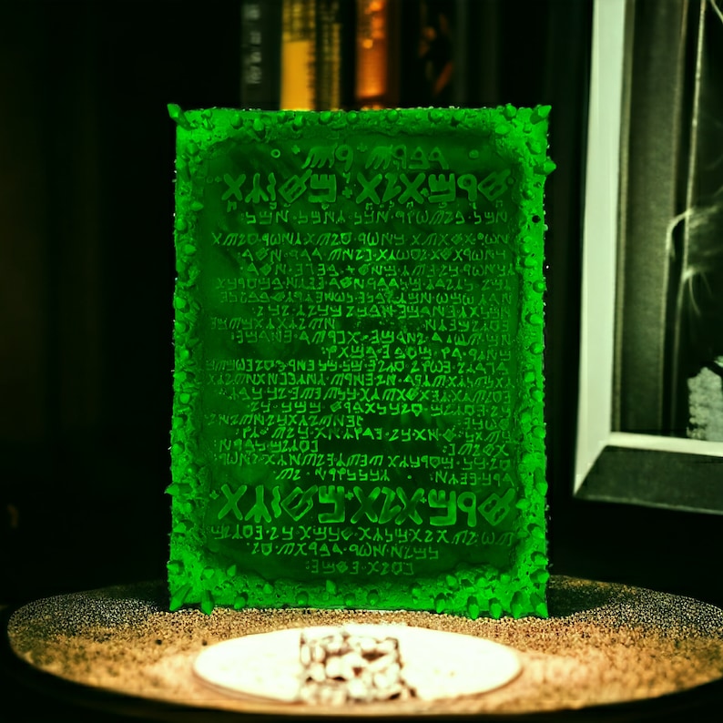 May include: A glowing green rectangular object with ancient-looking text etched across its surface. The edges have a textured, bubbly appearance. It sits on a round, golden-colored surface with a small, square object in front.