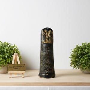 Puede incluir: Una réplica negra y dorada del Código de Hammurabi. La réplica presenta tallas detalladas en la parte superior y texto que cubre el cuerpo. El texto "CODE OF HAMMURABI REPLICA" está en un pequeño letrero frente a la réplica.