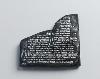 May include: A black, irregularly shaped stone plaque featuring a central engraved cross and extensive Latin text inscribed across its surface. The object has a weathered, dark texture with white-filled lettering that stands out against the dark base.