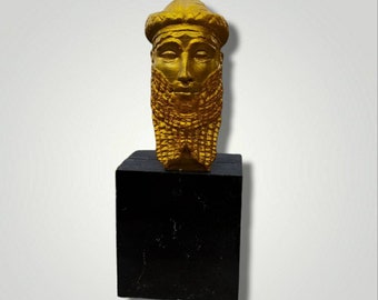 May include: A gold-colored bust of a bearded man with a crown on his head. The bust is on a black base.