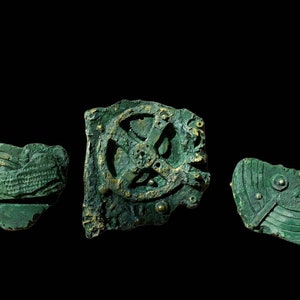 May include: Three antique bronze pieces with a green patina. The largest piece features a circular gear with a cross-shaped design. The other two pieces have geometric patterns.