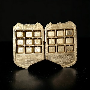 Puede incluir: Dos cajas de metal de color dorado con una cuadrícula de 16 cuadrados cada una. Cada cuadrado contiene un símbolo jeroglífico diferente. Las cajas están unidas por una bisagra y tienen un diseño decorativo en la parte delantera.