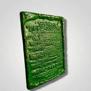 Op de afbeelding: Een groene, rechthoekige, getextureerde plaquette met een inscriptie in een oude taal. De plaquette heeft een ruw, onregelmatig oppervlak en lijkt van keramiek te zijn.