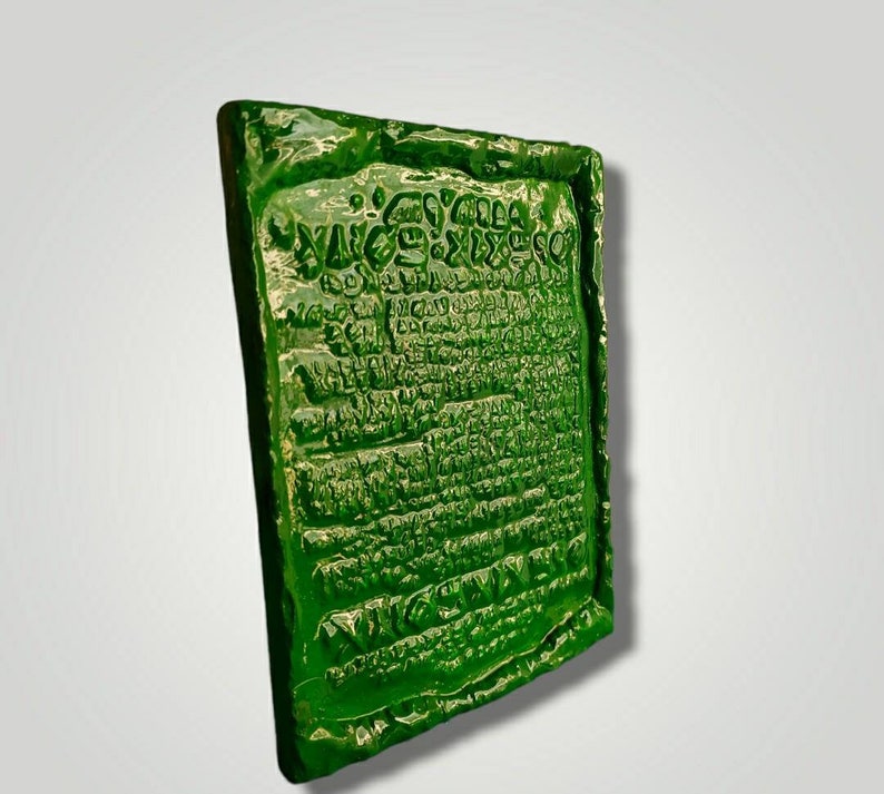 Op de afbeelding: Een groene, rechthoekige, getextureerde plaquette met een inscriptie in een oude taal. De plaquette heeft een ruw, onregelmatig oppervlak en lijkt van keramiek te zijn.