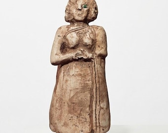 May include: A terracotta-colored clay figurine of a standing person with a textured, matte finish. The figure has short hair, a simple garment draped over the torso, and a single, prominent, bright green eye set into the face.