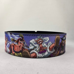 Puede incluir: Un cinturón de levantamiento de pesas negro con un diseño impreso colorido con personajes del anime One Piece. El diseño incluye un fondo morado con personajes en varias poses.