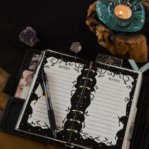 May include: A black and white planner with a starry night design and the word "NOTES" written on each page. A pen is lying on the open planner. There are crystals and a teal candle holder on a wooden surface.