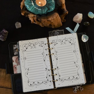 May include: A black leather bound planner with a white page open to a page with a decorative border. The border features a moon and stars. The planner is on a wooden surface with crystals and a teal glass candle holder.