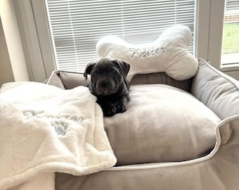 May include: A gray puppy rests in a plush dog bed with the name "Kaizer" embroidered on the side. A bone-shaped pillow and a soft blanket with the name "Kaizer" are also in the bed. The bed is placed near a window.