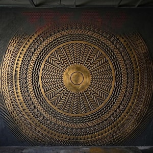 Può includere: Una grande opera d'arte murale circolare con un disco centrale dorato circondato da anelli concentrici di intricati motivi geometrici, tra cui zigzag, chevron e linee radianti su uno sfondo grigio scuro dalla superficie ruvida.