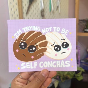 May include: A purple card with a white grid pattern and two cartoon concha pastries. The concha on the left is brown with white swirls and the concha on the right is white with a brown swirl. Both have big eyes and are crying. The text on the card reads "I'm trying not to be self conchas".