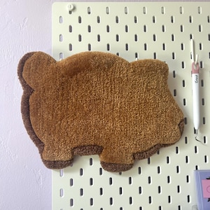 May include: A brown plush bear-shaped pillow with a brown border. The pillow is hanging on a white pegboard with holes.