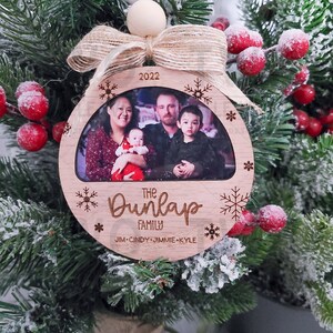 K&ouml;nnte beinhalten: Ein runder Weihnachtsbaumschmuck aus Holz mit einem Foto einer vierk&ouml;pfigen Familie. Der Schmuck ist mit Schneeflocken und dem Text "The Dunlap Family" und "Jim-Cindy-Jimmie-Kyle" verziert. Das Jahr 2022 ist ebenfalls auf dem Schmuckst&uuml;ck enthalten.