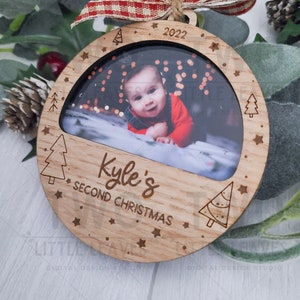 K&ouml;nnte beinhalten: Ein runder Weihnachtsbaumschmuck aus Holz mit einem Fotorahmen. Der Rahmen enth&auml;lt ein Foto eines kleinen Jungen. Der Schmuck ist mit Sternen und kleinen Weihnachtsb&auml;umen verziert. Der Text "Kyle's Second Christmas" und das Jahr "2022" sind in den Schmuck graviert.