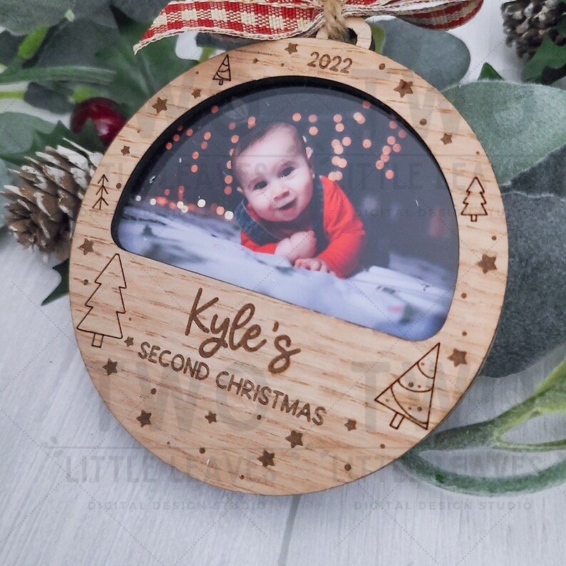 K&ouml;nnte beinhalten: Ein runder Weihnachtsbaumschmuck aus Holz mit einem Fotorahmen. Der Rahmen enth&auml;lt ein Foto eines kleinen Jungen. Der Schmuck ist mit Sternen und kleinen Weihnachtsb&auml;umen verziert. Der Text "Kyle's Second Christmas" und das Jahr "2022" sind in den Schmuck graviert.