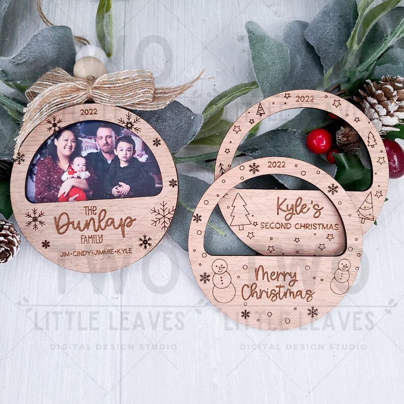 K&ouml;nnte beinhalten: Zwei h&ouml;lzerne Weihnachtsbaumschmuckst&uuml;cke mit einem Fotorahmen und einem Schneemann-Design. Das erste Ornament hat ein Familienfoto mit dem Text "The Dunlap Family" und "JIM-CINDY-JIMMIE-KYLE". Das zweite Ornament hat den Text "Kyle's Second Christmas" und "Merry Christmas".