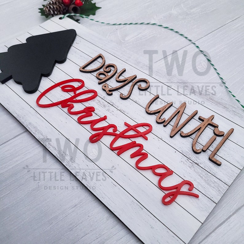 May include: White wooden sign with the words "Two Days Until Christmas" in red and brown lettering. The sign has a black wooden Christmas tree cutout at the top.