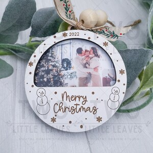 K&ouml;nnte beinhalten: Wei&szlig;er Weihnachtsbaumschmuck aus Holz mit einem Fotorahmen, einem Schneemann auf jeder Seite und dem Text "Frohe Weihnachten" in braunen Buchstaben. Das Jahr 2022 ist oben auf dem Ornament geschrieben.