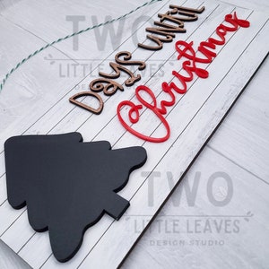 May include: A white wooden sign with the text "Days Until Christmas" in brown and red lettering. The sign has a black chalkboard tree shape in the bottom left corner. The sign also has the text "Little Leaves Design Studio" in the bottom right corner.