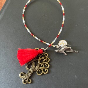 May include: A red tassel charm hangs from a beaded bracelet with a silver tree charm and a silver bird charm. The bracelet is made of white, gold, and red beads.