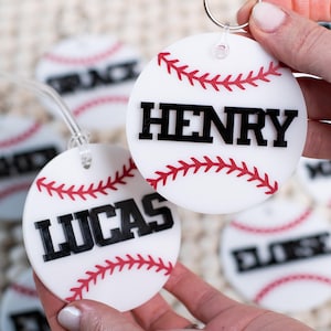 May include: White baseball-themed keychains with red stitching and black lettering. One keychain reads "HENRY", another reads "LUCAS". Other keychains with names are visible in the background. The keychains are round and have a silver key ring.