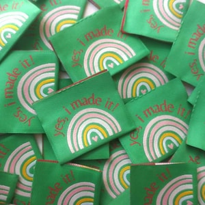 Puede incluir: Un montón de etiquetas de tela verde con un arcoíris y el texto "¡sí, lo hice!" en rojo.