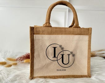 Può includere: Una borsa di juta marrone con un inserto in tela bianca. La borsa ha un design di due cerchi intrecciati con le lettere "I" e "U" all'interno, circondati da piccoli motivi floreali. La data "26.06.2099" è stampata sotto i cerchi.