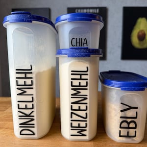 Può includere: Tre contenitori trasparenti in plastica per alimenti con coperchi blu. I contenitori sono etichettati con testo nero: DINKELMEHL, WEIZENMEHL e EBLY. Un contenitore più piccolo etichettato CHIA è posto sopra il contenitore WEIZENMEHL. I contenitori sono riempiti con ingredienti bianchi e beige.