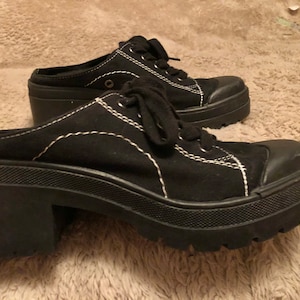 Puede incluir: Un par de zapatos de plataforma de lona negra con costuras blancas. Los zapatos tienen un tacón grueso y una punta redonda.