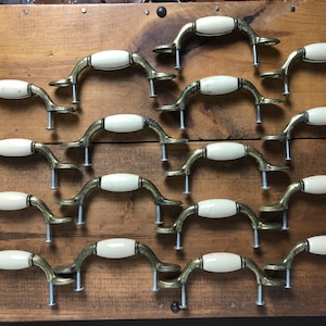 Puede incluir: Un juego de 18 pomos de armario vintage con una manija ovalada de cerámica blanca y una base de latón. Los pomos están dispuestos en filas sobre una superficie de madera.